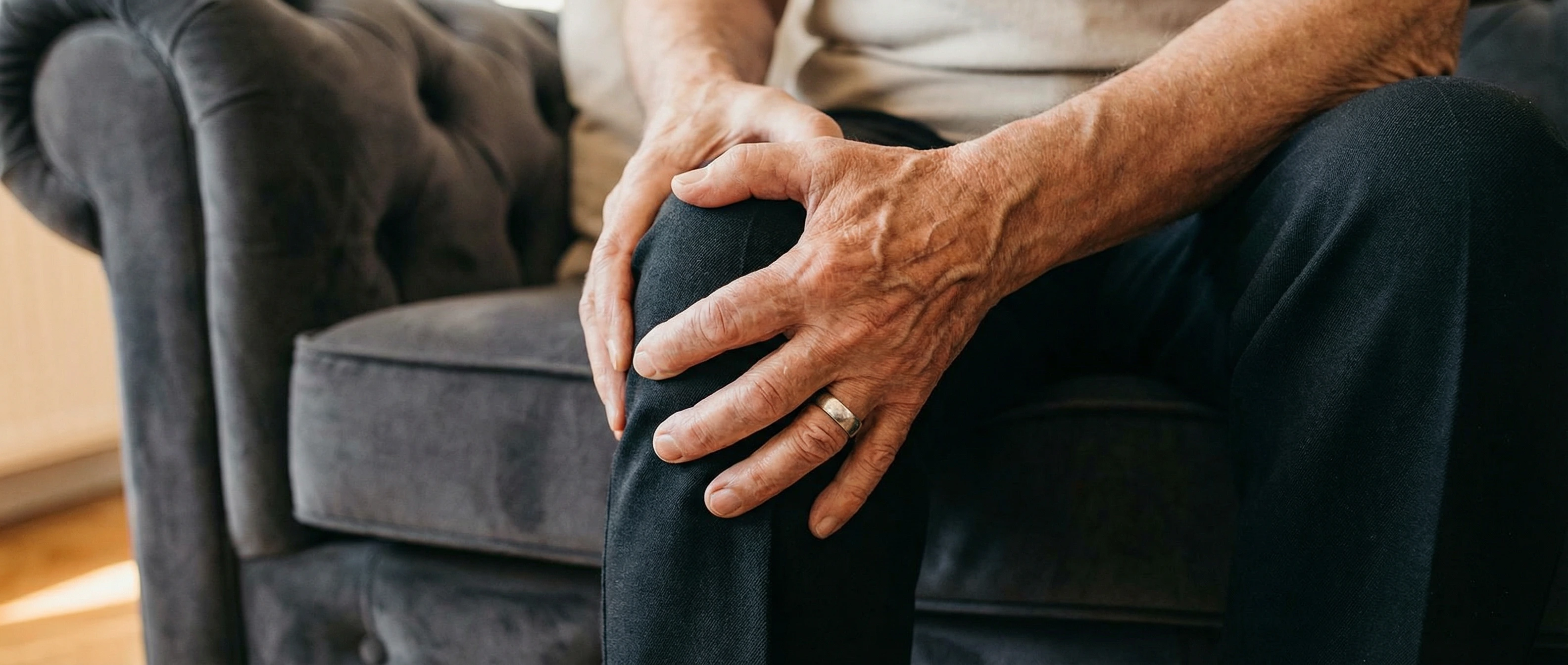 Elderly man holding his knee in pain while sitting on couch, experiencing chronic knee discomfort
