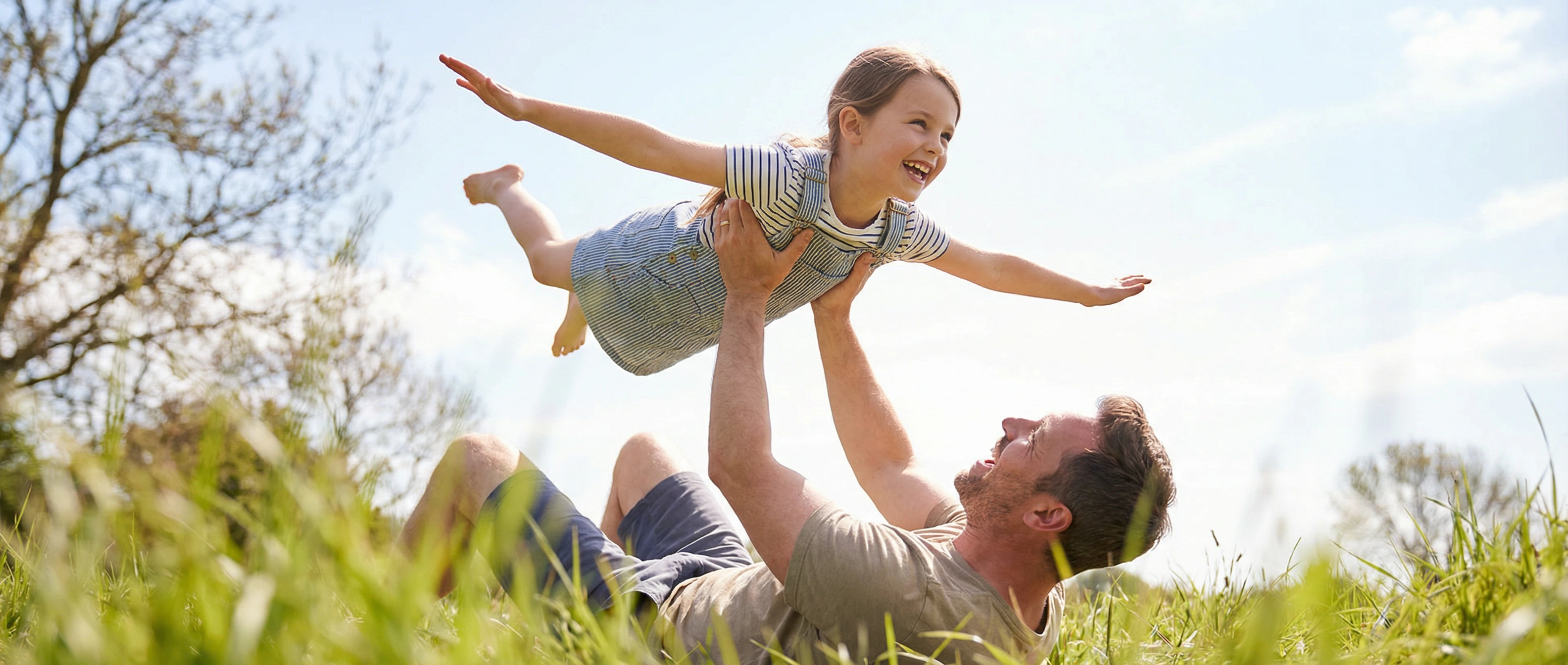 Father lying in grass lifting his daughter in the air, enjoying pain-free shoulder mobility after treatment